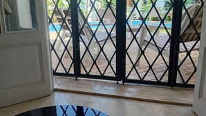 On aperçoit, à travers une porte grillagée, un espace extérieur avec une piscine, des chaises longues et des plantes tropicales.