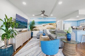 A modern living room with a beach mural, a grey sofa, a coffee table, and a blue armchair. - Gulf View • 199 Steps to Beach • Dog-Friendly (Indian Rocks Beach)
