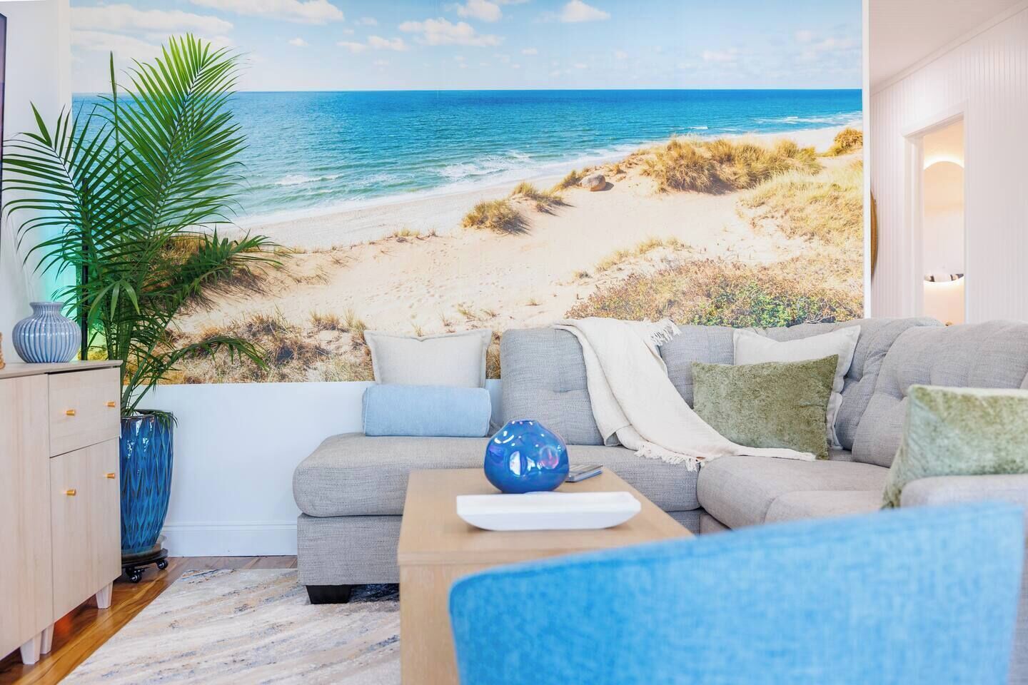 Un salon avec une grande fresque murale représentant une plage, un canapé gris et un vase bleu.