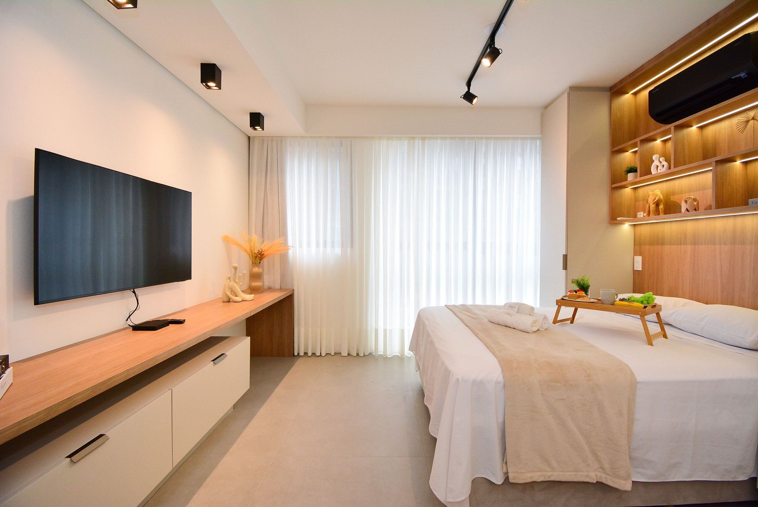 Una habitación de hotel moderna con televisor de pantalla plana, una mesita consola de madera, una cama con colcha blanca y una mesita de noche con bandeja.