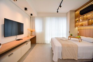 A modern hotel room with a flat-screen TV, a wooden console table, a bed with a white bedspread, and a bedside table with a tray. - Elegante Studio frente a UFSC e HU -201R (Florianópolis)