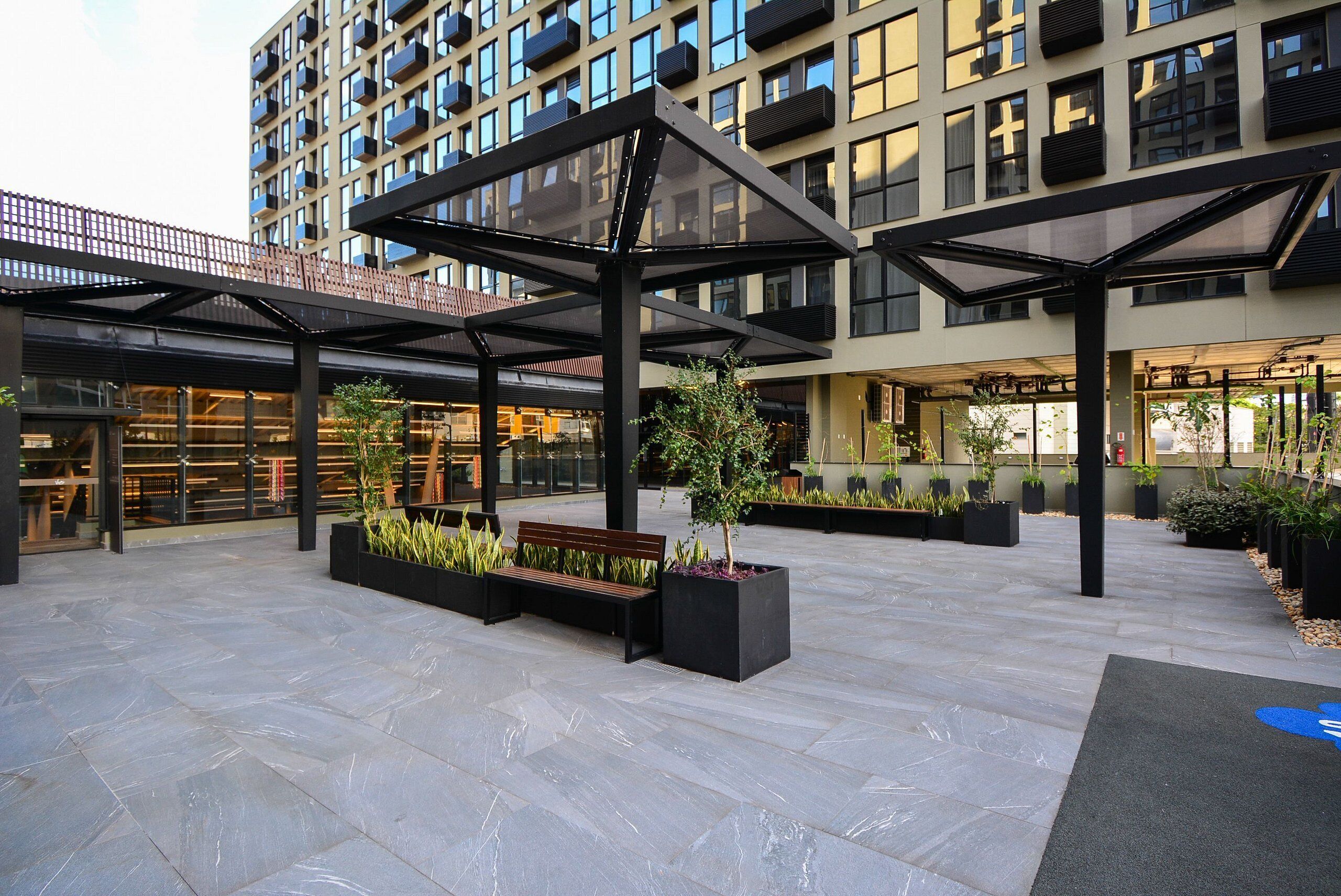 A modern outdoor area with a covered seating area, planters with greenery, and a building with multiple balconies.