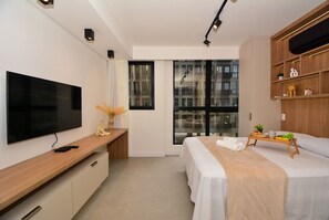 Habitación de hotel moderna con televisión de pantalla plana, una mesa consola de madera, una cama con colcha color beige, una mesita de noche con una bandeja de fruta y un ventanal con vistas a edificios.