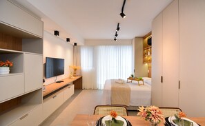 A modern hotel room with a flat-screen TV, a bed, a dining area with a table and chairs, and a wardrobe. - Elegante Studio frente a UFSC e HU -201R (Florianópolis)