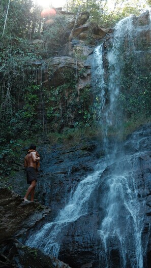 A waterfall in a forested area with a person standing at the base. - Canto dos Pireneus Hospedaria (Pirenopolis)