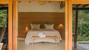 A bedroom with a bed, bedside lamps, a bench with towels, and a view of greenery. - Canto dos Pireneus Hospedaria (Pirenopolis)