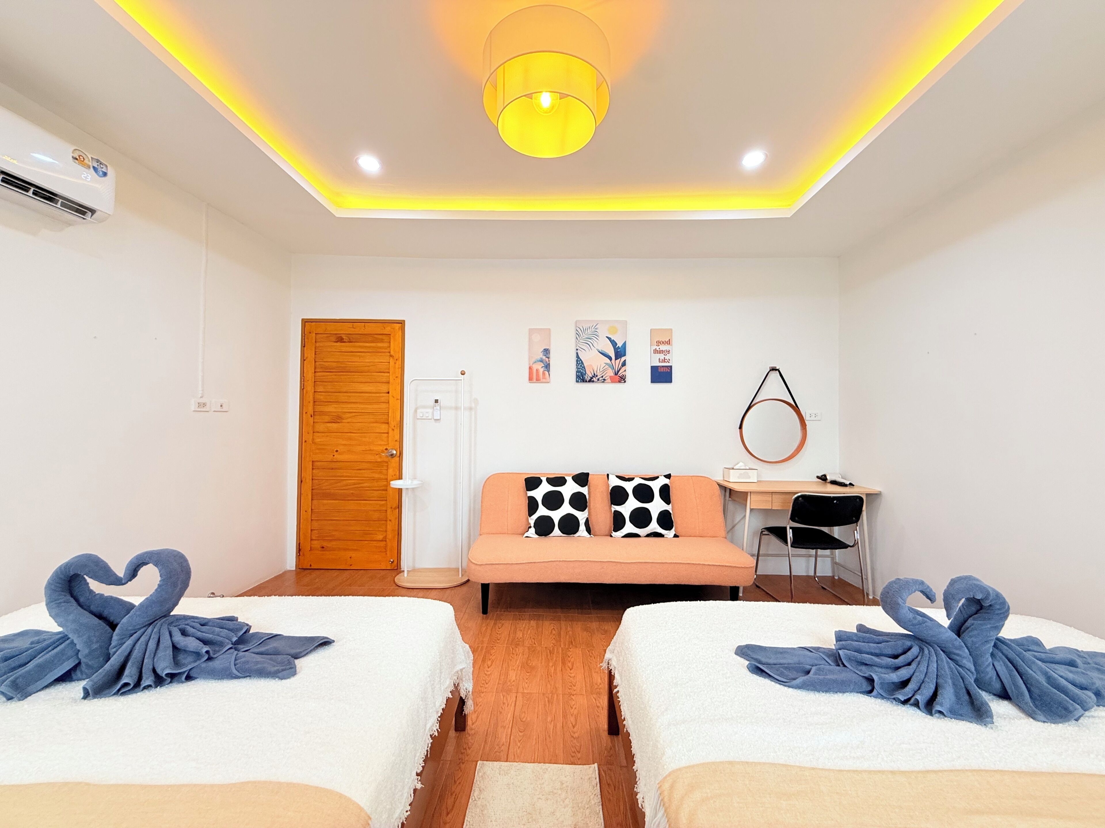 A hotel room with two beds, a wooden door, a desk, a chair, a wall-mounted mirror, and a ceiling light fixture.