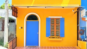 Ein gelbes Gebäude mit blauer Tür und Fensterläden, einer gefliesten Eingangszone und einem kleinen Blumenkasten.