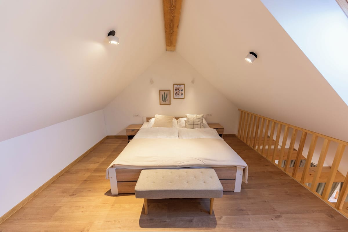 A bedroom with a large bed, a bench, wooden flooring, a sloped ceiling, and a wooden railing.