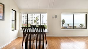 Een eetgedeelte met een tafel en stoelen, grote ramen met uitzicht op gebouwen en de zee.