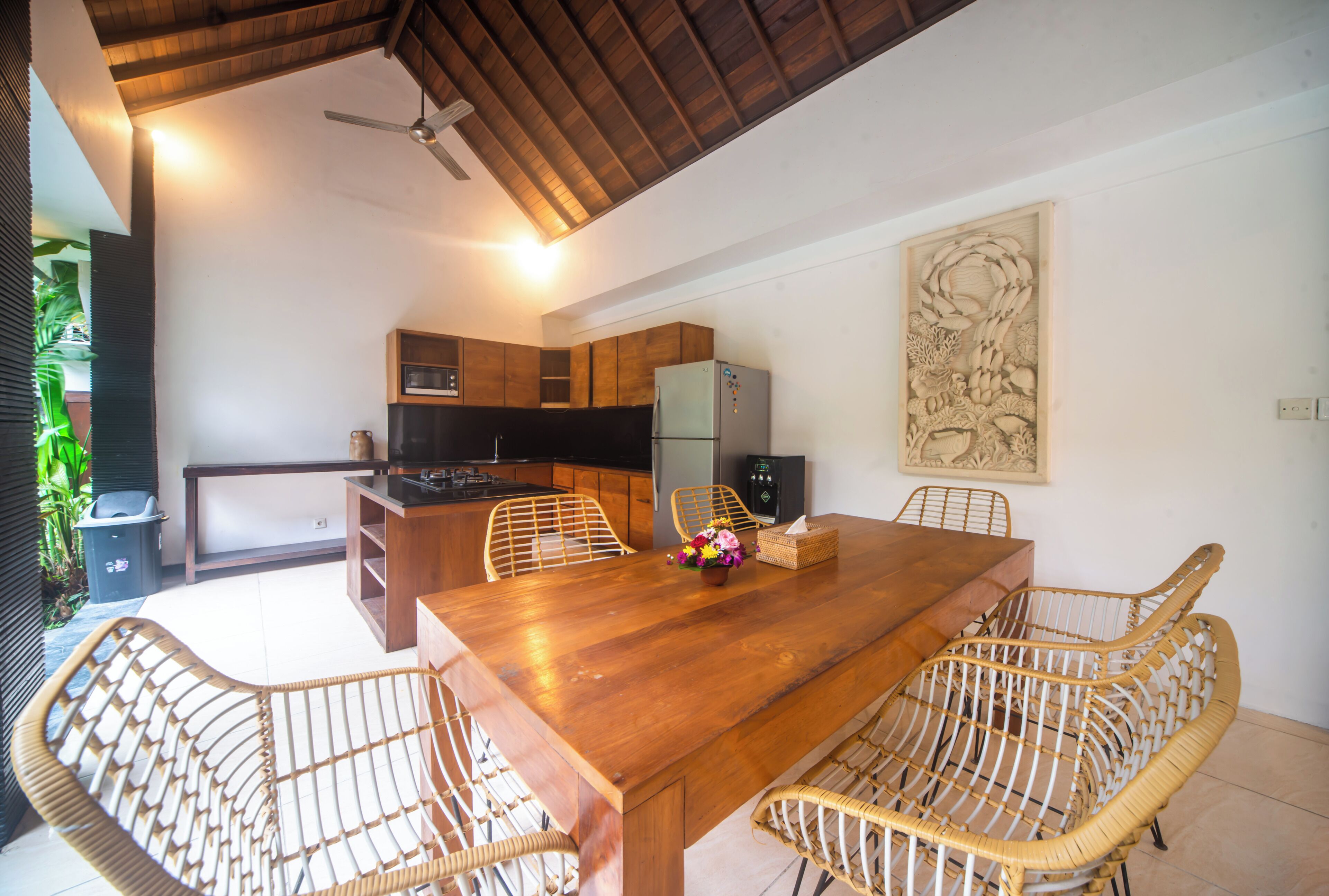 A dining area with a wooden table and wicker chairs, a kitchen with wooden cabinets and a refrigerator, and a ceiling fan.