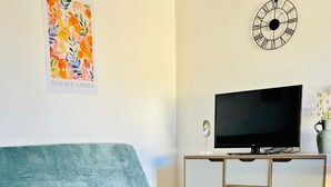 En stue med en turkis sofa, et fjernsyn på et træskab, et vægur og en skål med frugt på et lille bord.