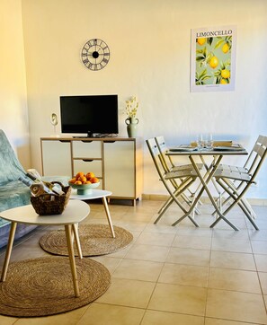 A living room with a sofa, TV, dining table, chairs, and a wall clock. - Waterfront studio sleeps 4 (Santa-Maria-Poggio)