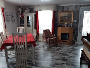 A dining area with a red tablecloth, white chairs, a wooden cabinet with a painting, and a piano.