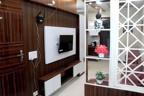 A modern living room with a flat-screen TV mounted on a wooden wall, a white entertainment unit below, and a decorative white partition with a geometric pattern. - The Soft Palace 3BHK near Dwarka (Delhi)