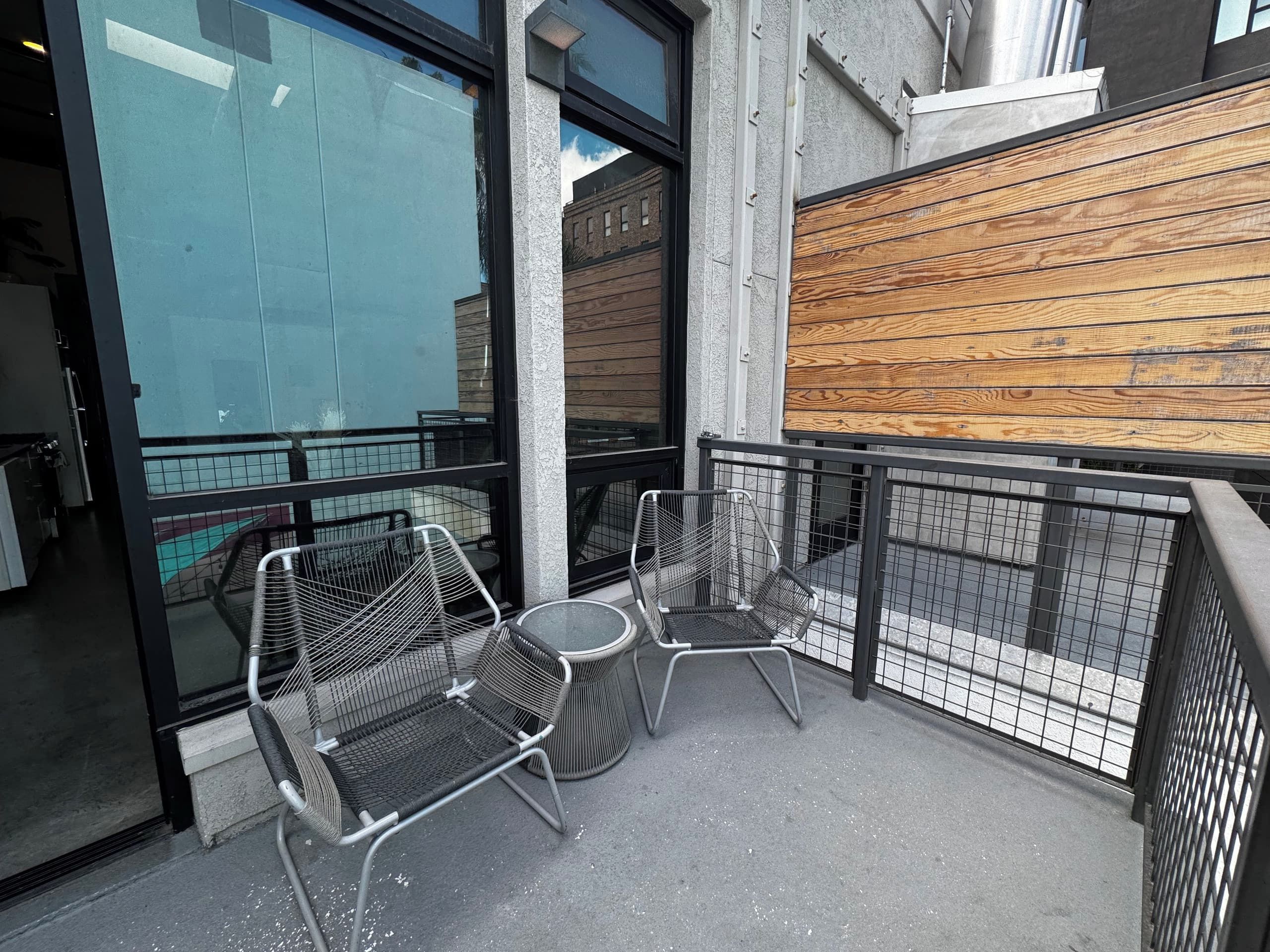 A balcony with metal chairs and a small table, a wooden wall, and a glass door reflecting the outside.