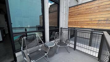 A balcony with metal chairs and a small table, a wooden wall, and a glass door reflecting the outside.