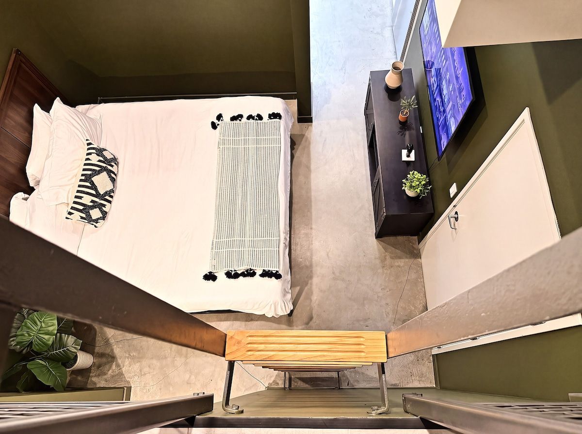 A staircase with a wooden handrail, a bed with a white and black patterned blanket, and a wall-mounted TV.