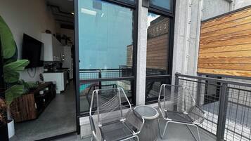 Un balcón con dos sillas de metal y una mesita redonda, una zona de cocina con un televisor, y vista a un edificio a través del ventanal.