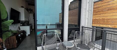 A balcony with two metal chairs and a small round table, a kitchen area with a TV, and a view of a building through the glass door.