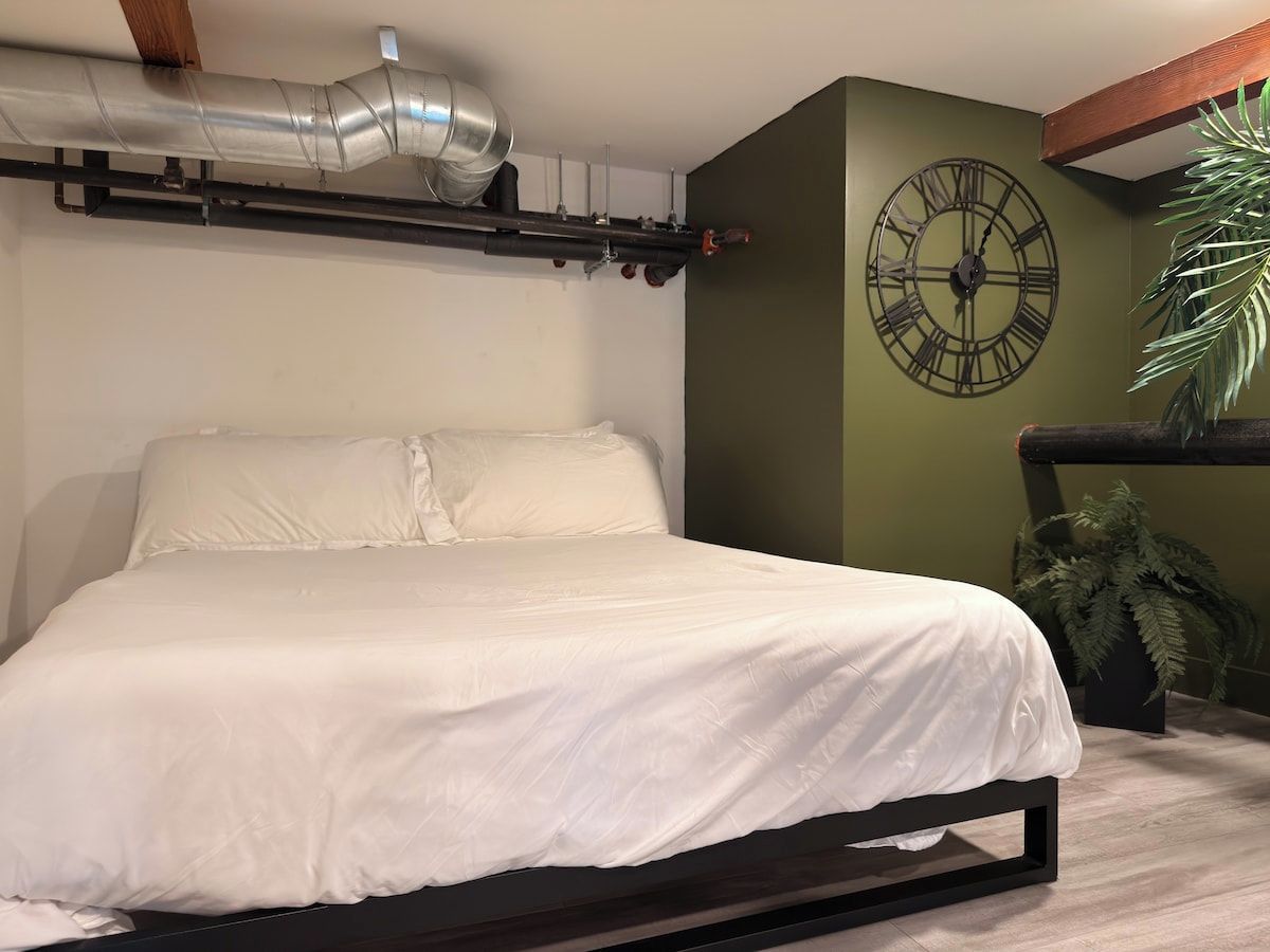 Un lit avec un cadre noir, un matelas blanc et une tête de lit adossée à un mur vert. On y voit une grande horloge murale avec des chiffres romains.