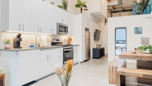 A modern kitchen with white cabinets, stainless steel appliances, and a dining area with a wooden table and bench. - Stylish Downtown Loft Near Disney (Santa Ana)