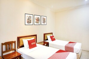 Une chambre d’hôtel avec deux lits, chacun doté d’une tête de lit en bois et d’un oreiller rouge orné du logo « OYO ». On peut voir des œuvres d’art représentant des feuilles, encadrées, accrochées au mur, ainsi qu’une table de chevet et une chaise en bois.