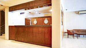 Un mostrador de recepción con un letrero que dice 'CAJERO', dos relojes, una escalera y una zona de estar con sillas a rayas y un sofá.