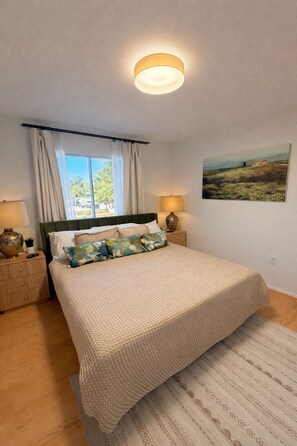 Ein Schlafzimmer mit einem Bett, einem Nachttisch, einer Lampe, einem Fenster mit Vorhängen und einem Bild an der Wand.