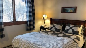 A bedroom with a bed, a nightstand, a lamp, a window with curtains, and a painting on the wall. - Cabin close to hiking trails and ski resorts (South Lake Tahoe)