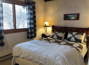 Ein Schlafzimmer mit Bett, Nachttisch, Lampe, einem Fenster mit Vorhängen und einem Bild an der Wand.