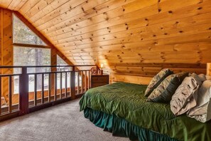 Un dormitorio con techo inclinado, una cama con ropa de cama verde, una mesita de noche y una ventana con vistas a un bosque.