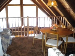 A cozy attic room with a wooden dining table and chairs, a white cushioned bench, and a view through a barred window.