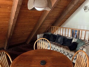 Ein Holzesstisch mit Stühlen, eine Hängelampe und eine Couch mit gemusterten Kissen in einem Raum mit freiliegenden Holzbalken.
