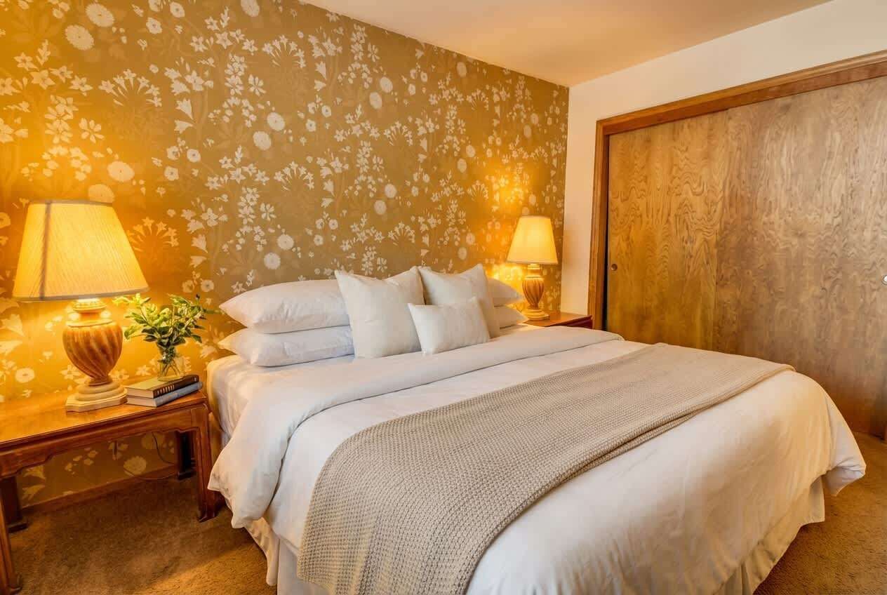 Een net opgemaakt bed met witte lakens, twee bedlampen, een houten nachtkastje en een behang met een bloemenmotief.