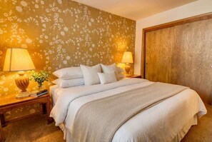 A neatly made bed with white linens, two bedside lamps, a wooden nightstand, and a floral wallpaper pattern. - Tahoe Cabin with Stone Fireplace, 2-Car Garage & Fenced Yard — Steps to the River (South Lake Tahoe)