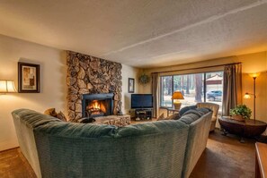 A living room with a stone fireplace, a television, a sofa, and a view of the outside. - Tahoe Cabin with Stone Fireplace, 2-Car Garage & Fenced Yard — Steps to the River (South Lake Tahoe)