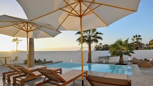 Zona de piscina con sillones de descanso y sombrillas, una piscina, palmeras y vista al atardecer.
