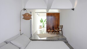 Una habitación moderna con una cama, una mesita y una silla. Se ve una planta y una puerta de madera al fondo.