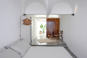 Ein modernes Zimmer mit einem Bett, einem kleinen Tisch und einem Sessel. Im Hintergrund befindet sich eine Pflanze und eine Holztür.