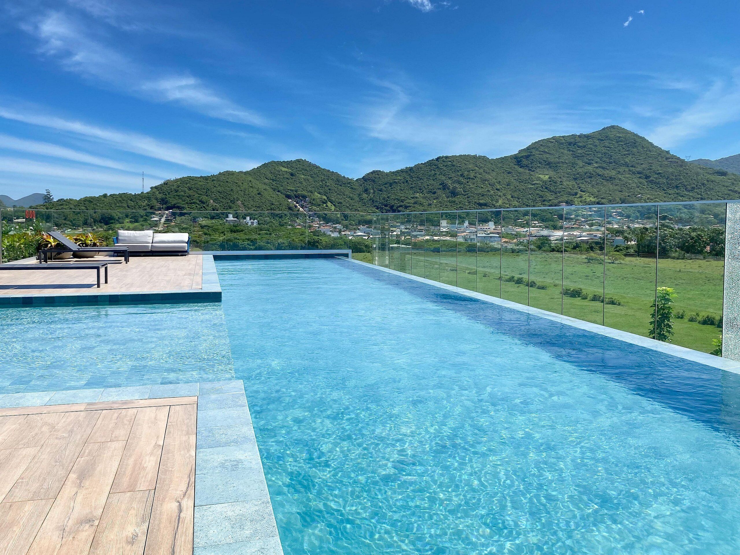 Una piscina con muelle de madera, sillas de descanso y vistas a colinas verdes y edificios.