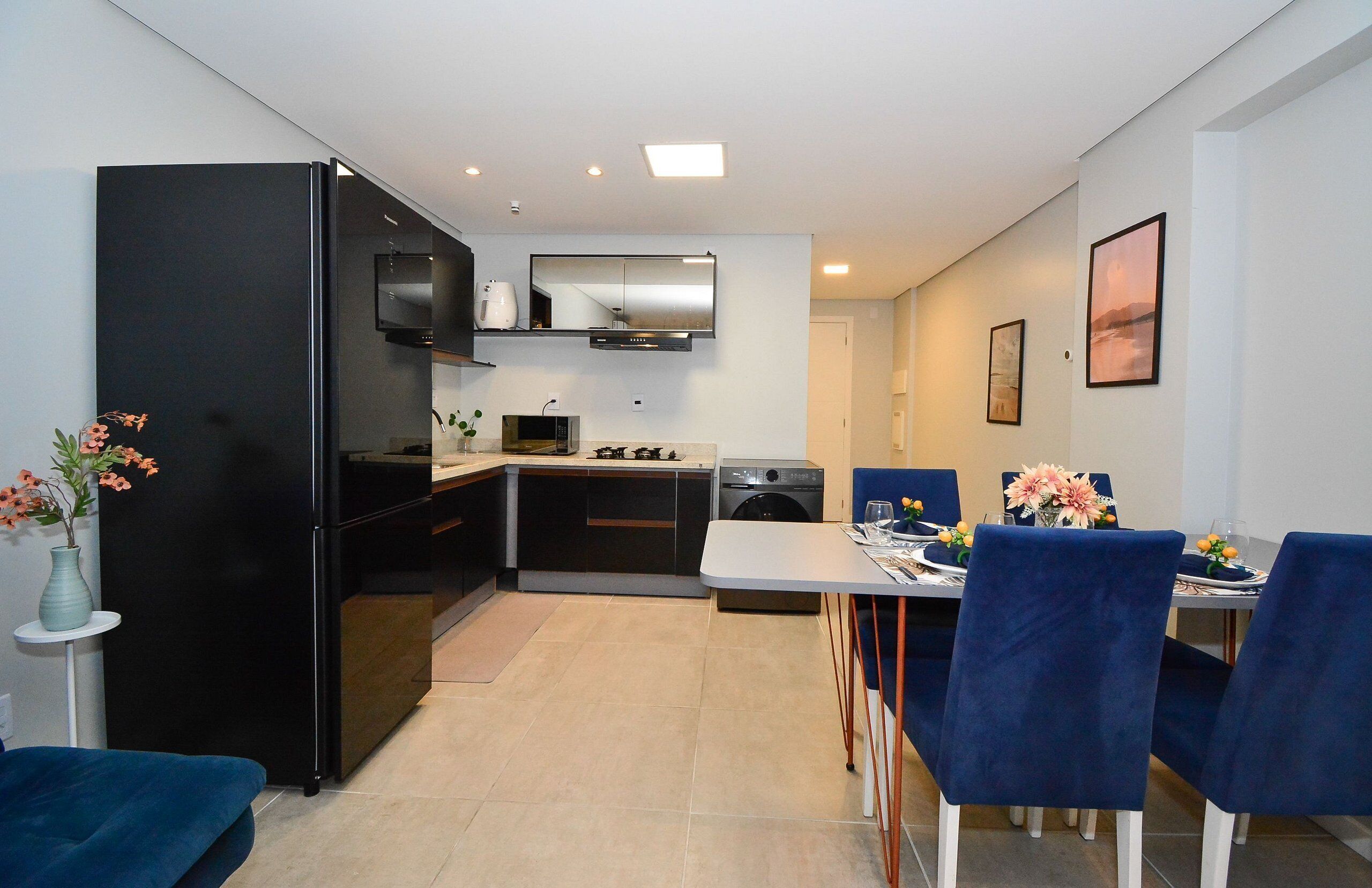 Una cocina moderna con un refrigerador, microondas y horno negros, una mesa de comedor con sillas azules y una pequeña mesita con un florero.