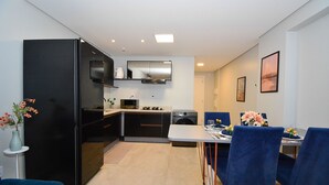 Una cocina moderna con un refrigerador, microondas y horno negros, una mesa de comedor con sillas azules y una pequeña mesita con un florero.