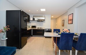A modern kitchen with a black refrigerator, microwave, and oven, a dining table with blue chairs, and a small side table with a vase. - Studio Shopping OKA Floripa - SOK239 (Florianópolis)