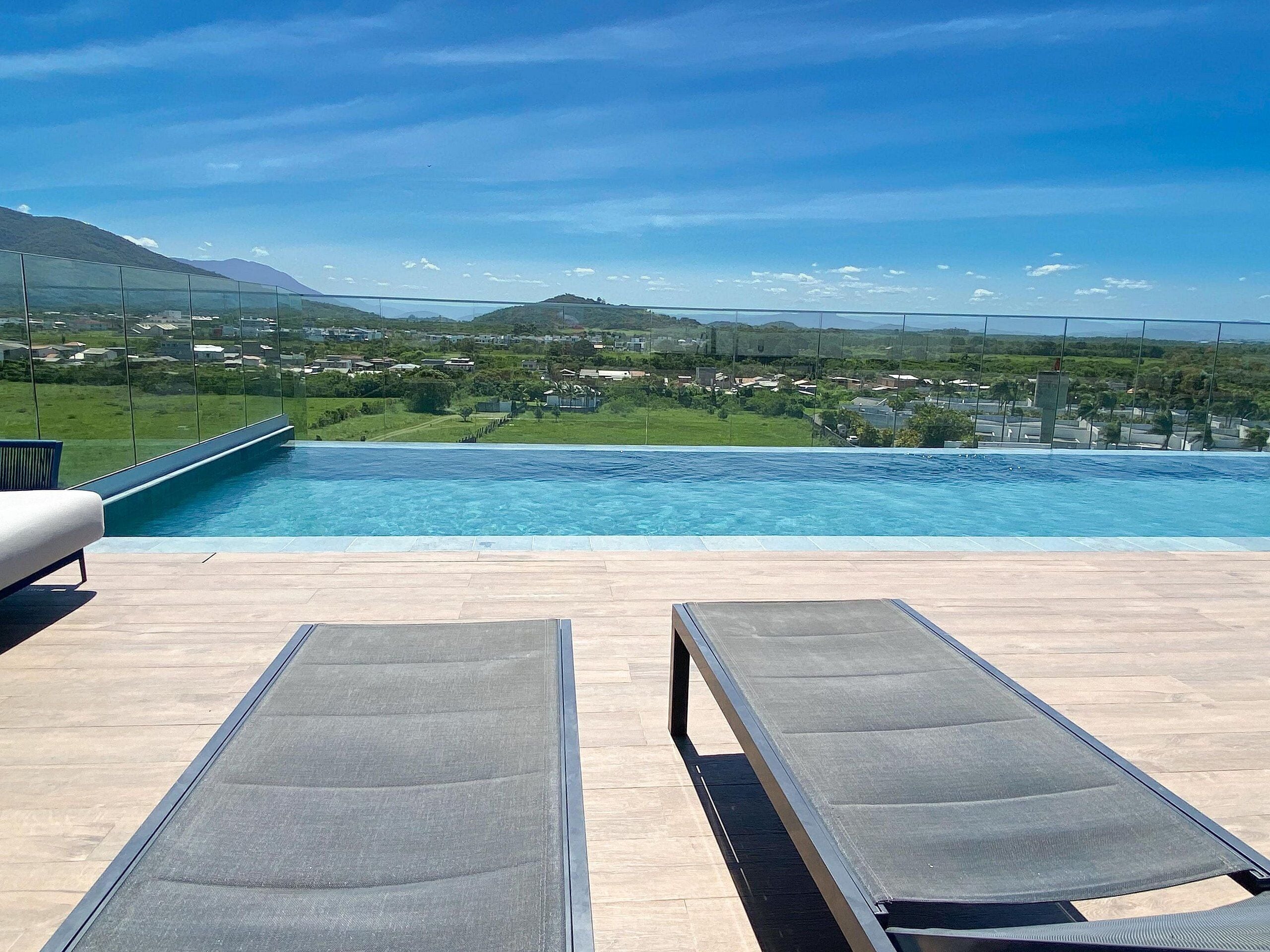 Vista da piscina com duas espreguiçadeiras, céu claro e paisagem distante.