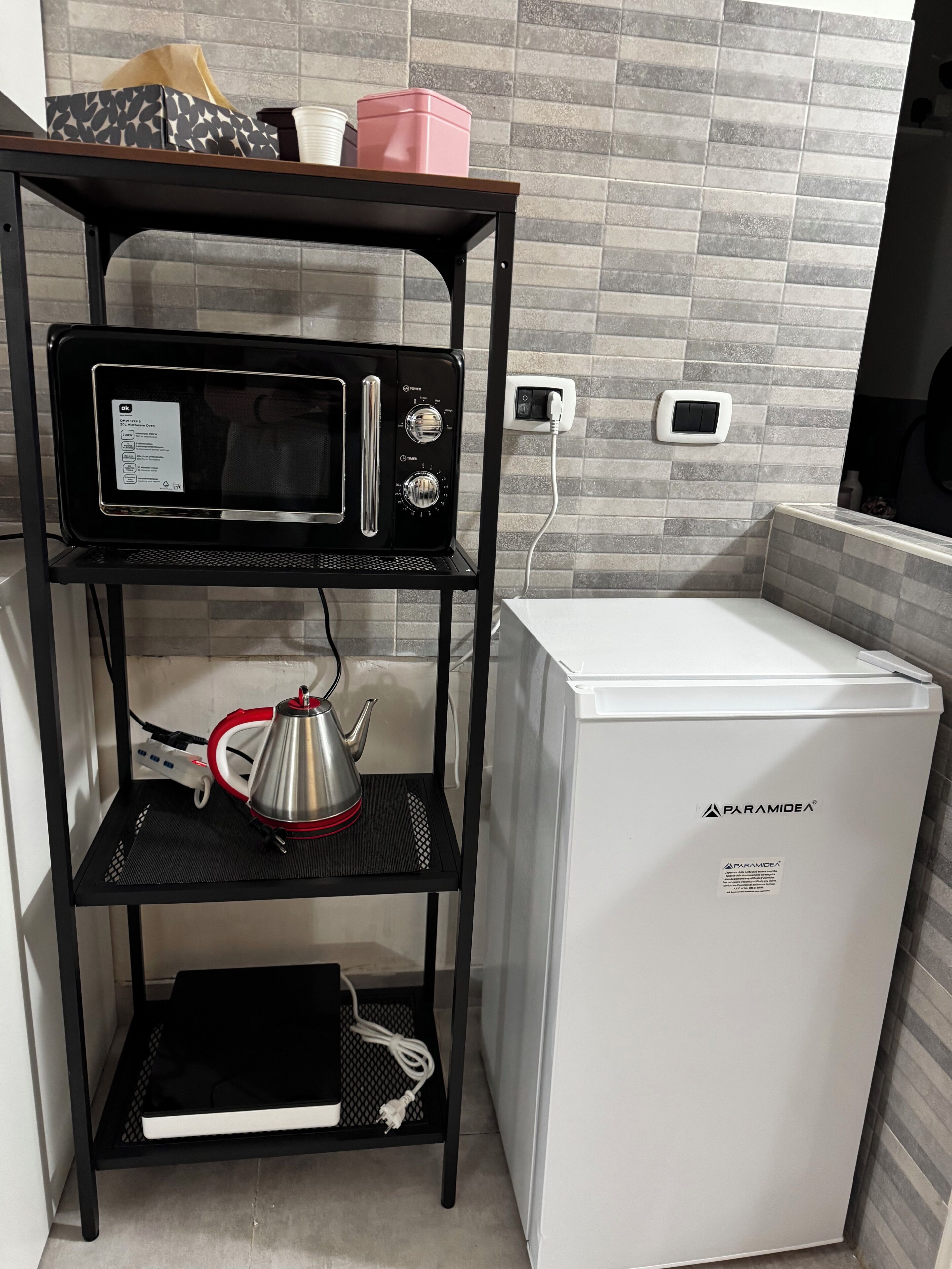 A compact kitchenette with a microwave, a small refrigerator, and a shelf unit with a kettle and a power strip.