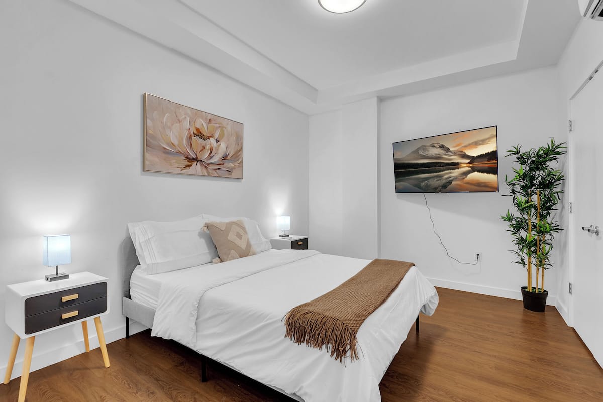 A modern bedroom with a bed, bedside tables, a lamp, a painting, a TV, and a potted plant.