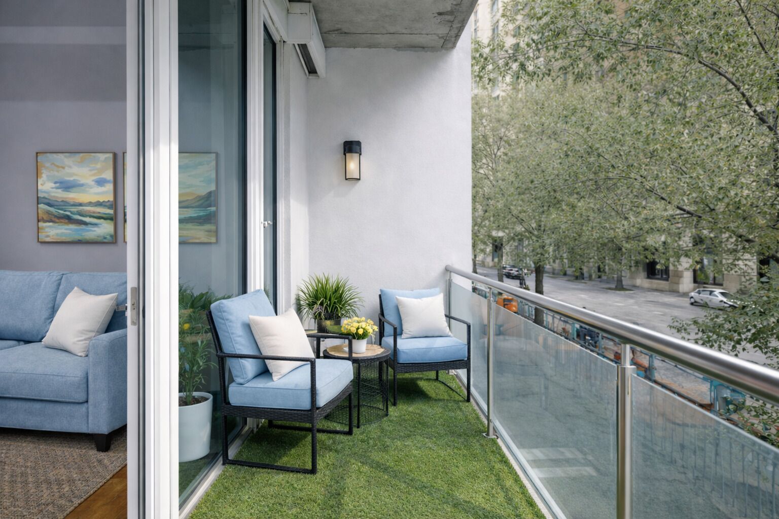 Un balcon avec deux chaises bleues, une petite table ornée d’une plante et une balustrade en verre donnant sur une rue.