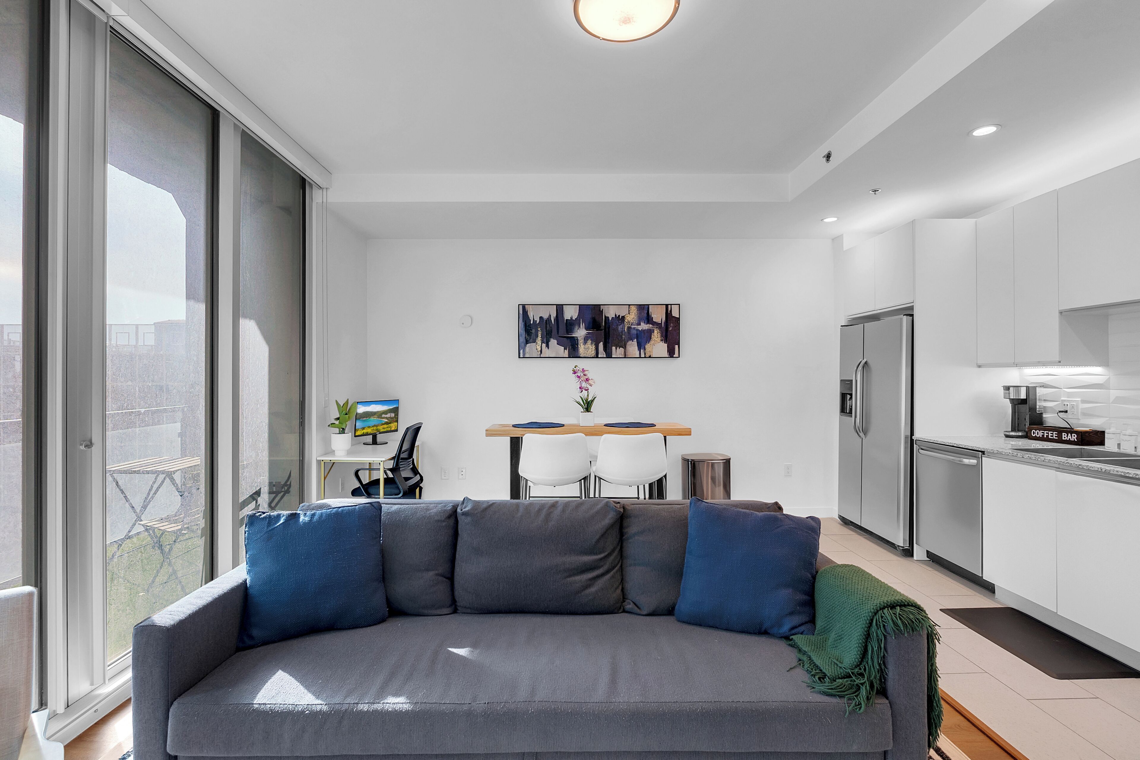 Un salon moderne avec un canapé gris, une table à manger avec des chaises, un coin cuisine avec un réfrigérateur et une grande fenêtre.
