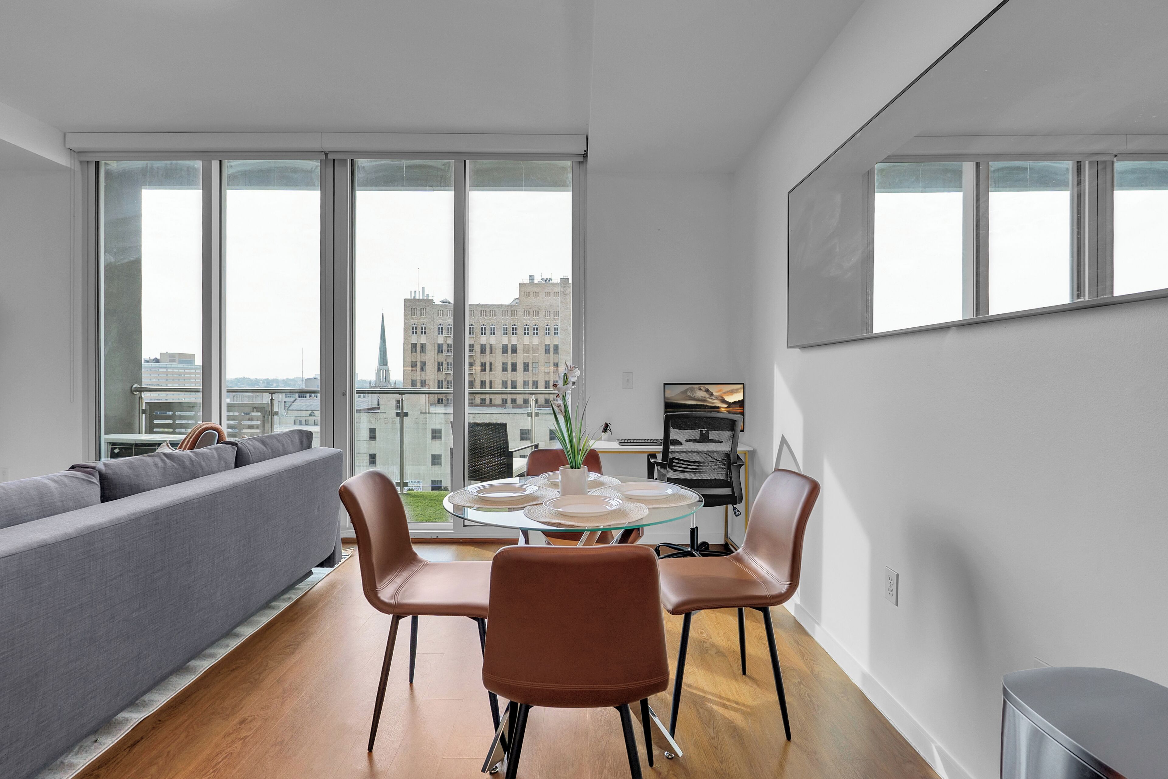 A modern living room with a sofa, dining table, chairs, and large windows offering a city view.